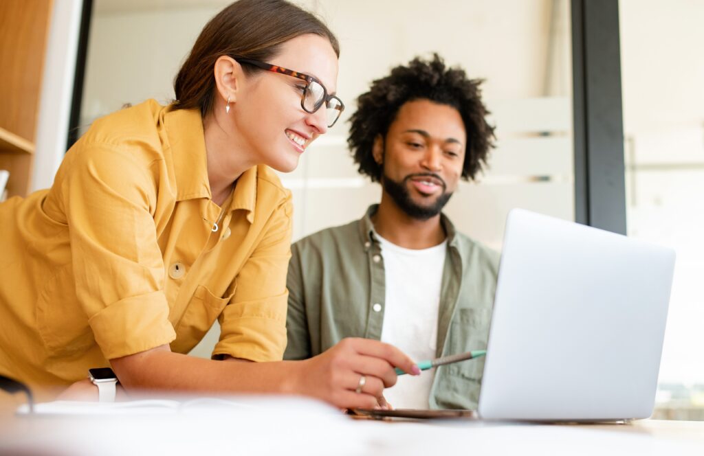 Two colleagues have pleasant conversation sitting side by side in modern open space office in front of laptop, smiling and enjoying friendly working atmosphere, brainstorming together, solving tasks.